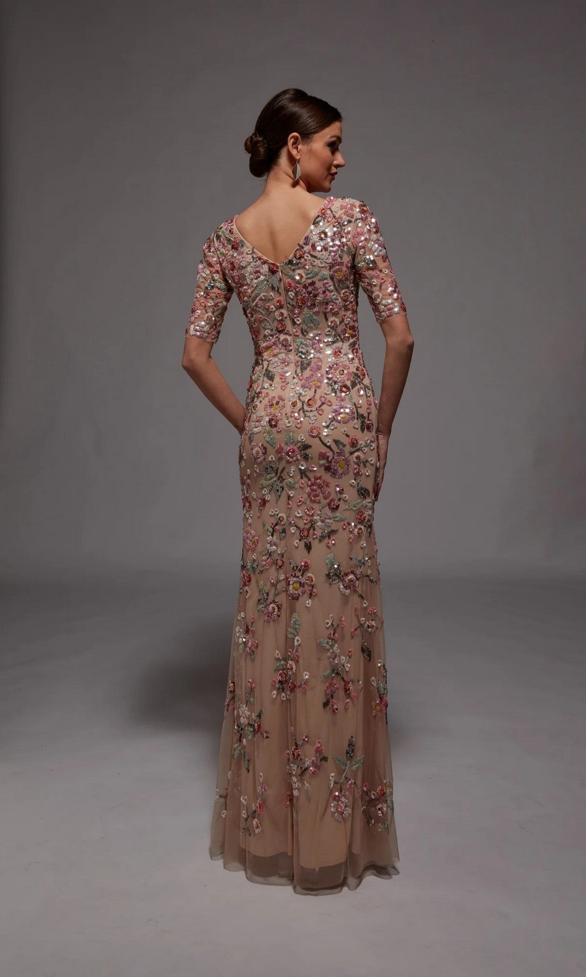 A woman faces away, modeling the Long Formal Dress: Alyce 27848—a fitted, floor-length nude gown with short sleeves and floral beaded appliques—perfect for formal occasions, set against a plain gray background.